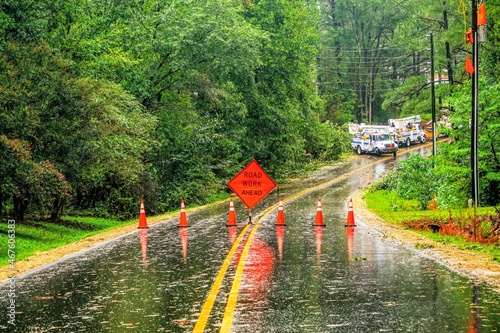 A view of an emergency roadblock during a storm on a rural road with orange cones and utility trucks in the background.