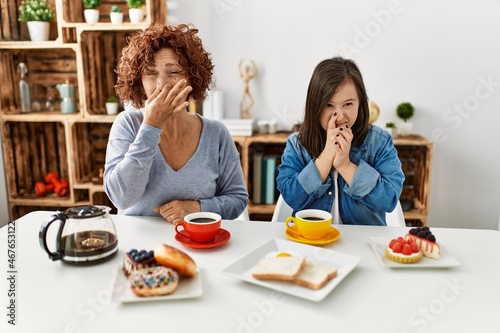 Family of mother and down syndrome daughter sitting at home eating breakfast smelling something stinky and disgusting, intolerable smell, holding breath with fingers on nose. bad smell
