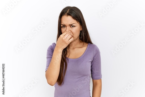 Young brunette woman standing by isolated background smelling something stinky and disgusting, intolerable smell, holding breath with fingers on nose. bad smell