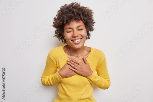 Pleased woman with curly hair sincere smile on face presses hands to chest feels thankful for help closes eyes wears casual yellow jumper poses against white background flattered of smth heartwarming