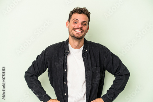Young caucasian man isolated on green background confident keeping hands on hips.