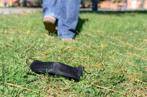 Some feet walk away and leave behind a black surgical mask on the ground on the grass. Concept of end of use due to the pandemic caused by covid19.