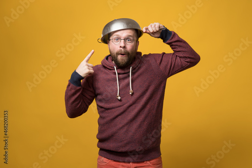Pastafarianism. Cute man puts a colander on head and points at it with finger. Parody of religion. Studio, yellow background.