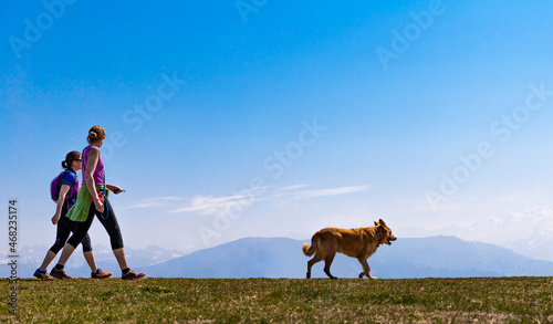 Two sportive women with a dog are walking with mountains and blue sky as background