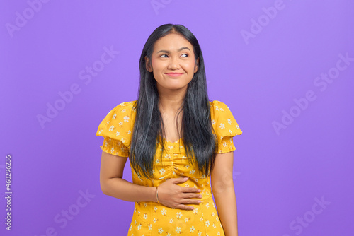 Young Asian woman keep hands on stomach and feel satiety after eating tasty on purple background