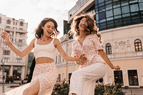 Beaming lovely caucasian young women dancing vigorously outdoors in city center. Magical teenagers with flying hair hold hands, dressed in white clothes. Lifestyle, female beauty concept