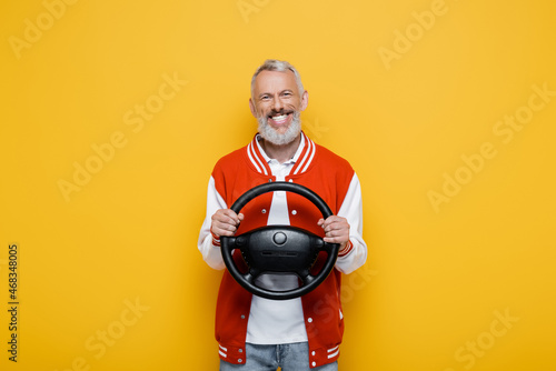 happy middle aged man in bomber jacket holding steering wheel isolated on yellow