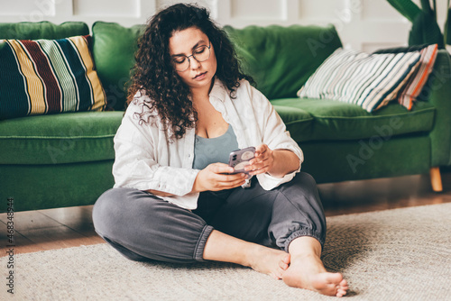 Curly haired plus size young woman wearing comfortable clothes in glasses clicks on smartphone screen sitting on floor mat in stylish room