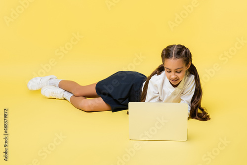 Schoolkid in shirt using laptop while lying on yellow background