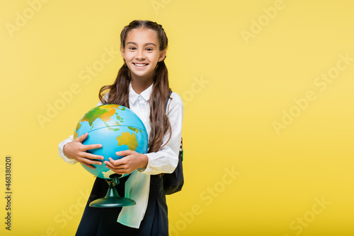 Happy schoolgirl holding globe and looking at camera isolated on yellow
