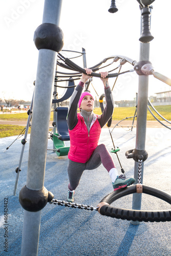 Young caucasian fit woman in warm pink fitness wear stretching her leg with hand on bar of modern athletic field after training outdoors on sunrise on autumn, winter or spring morning. Sports, active