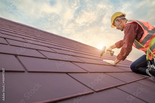 Roofer working in special protective work wear gloves, using air or pneumatic nail gun installing concrete or CPAC cement roofing tiles on top of the new roof under construction residential building