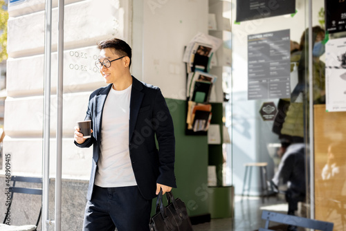 Asian businessman with coffee going from entrance of building. Smiling modern adult successful man wearing suit and glasses with briefcase. City at sunny day