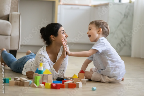 Happy nanny giving high five praise to excited preschool kid boy for completing toy tower on warm floor. Indian babysitter and kid playing at home, constructing building from small wooden blocks