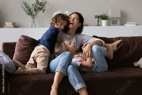 Overjoyed mother with two kids having fun on comfortable couch at home, laughing happy mom with adorable daughter and son tickling, engaged in funny active game, spending leisure time together