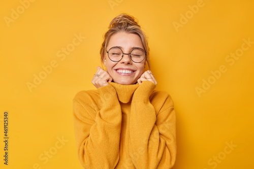 Cheerful young woman enjoys pleasant memories wears soft comfortable jumper and round spectacles keeps eyes closed smiles pleasantly isolated over yellow background recalls nice moment in life