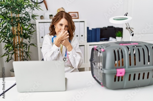 Middle age veterinarian woman working at pet clinic smelling something stinky and disgusting, intolerable smell, holding breath with fingers on nose. bad smell