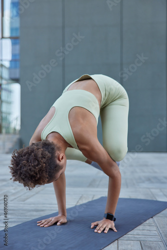 Vertical shot of slim curly haired woman practices advanced yoga leans downwards has athletic strong body pose on fitness mat dressed in activewear against grey wall does exercises outdoors.