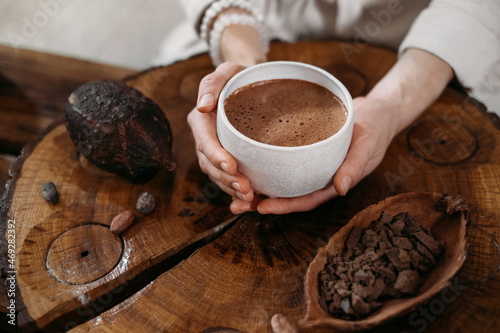 Hot handmade ceremonial cacao in white cup. Woman hands holding craft cocoa, top view on wooden table. Organic healthy chocolate drink prepared from beans, no sugar. Giving cup on ceremony, cozy cafe