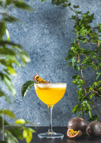 Glass of refreshing passionfruit Martini cocktail served on gray table surface surround of fresh passion fruit and different green plants, shallow depth of the field.
