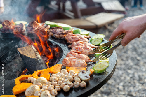 Close-up detail view of chef flipping by tongs tasty crispy marinated quails and vegetables grilled at round steel iron firepit hearth table surface brazier with burning firewoods. Barbecue yard home