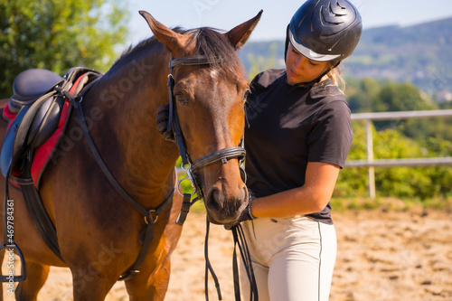 Posing of a Caucasian blonde girl on a horse caressing and pampering a brown horse, dressed in black rider with safety cap