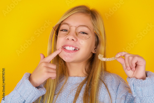 caucasian kid girl wearing blue knitted sweater over yellow background holding an invisible aligner and pointing to her perfect straight teeth. Dental healthcare and confidence concept.