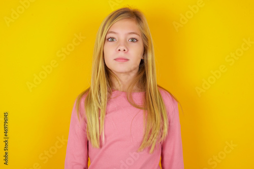 Stunned caucasian little kid girl wearing long sleeve shirt over yellow background stares reacts on shocking news. Astonished blonde kid girl holds breath