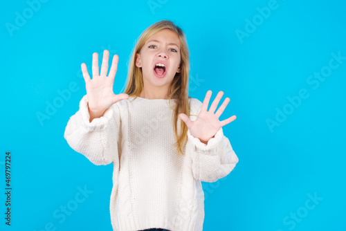 Dissatisfied caucasian little kid girl wearing wool jersey over blue background frowns face, has disgusting expression, shows tongue, expresses non compliance, irritated with somebody.