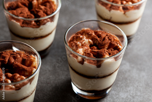 Delicious traditional tiramisu desert served in small glass cups on stone countertop. Low light image shows texture and layers from angle with shallow depth of field.