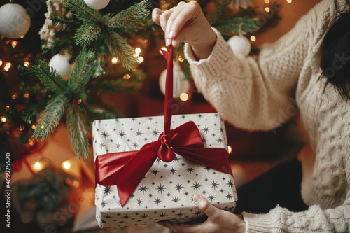 Hands in cozy sweater opening christmas gift with red bow on background of christmas tree with lights. Stylish female holding present with red ribbon in festive room close up. Merry Christmas!