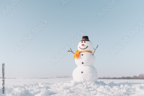 Funny snowman in stylish brown hat and yellow scalf on snowy field. Blue sky on background