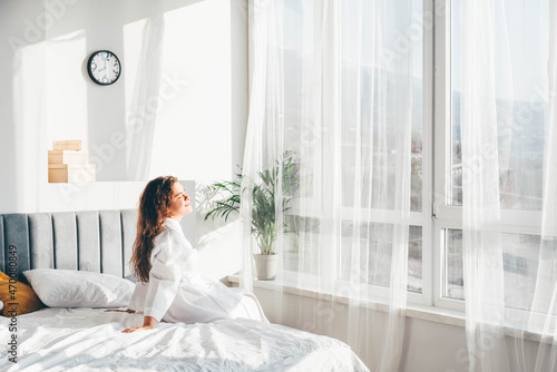  Woman in white bathrobe sitting near the big white window on bed after waking up.