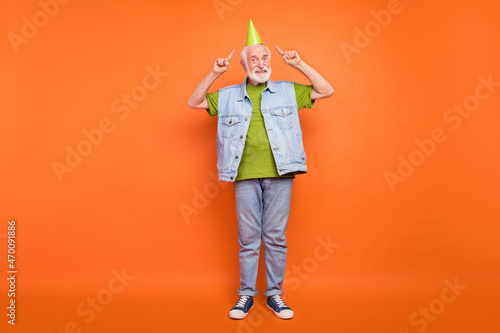 Photo of pretty handsome man pensioner dressed retro denim vest pointing fingers anniversary cap isolated orange color background