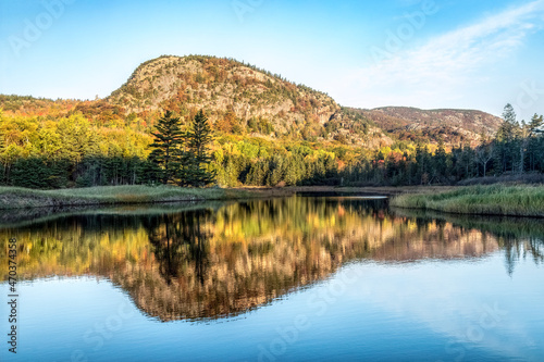 The Beehive, a rocky summit on Mt. Desert Island in Acadia National Park, Maine, is reflected upon Beehive Lagoon behind Sand Beach surrounded by trees with fall foliage color.