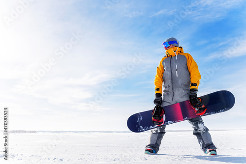 Young man holds a snowboard in his hands against the background of a winter landscape. Male in ski goggles and overalls with his board on white snow on a sunny frosty day go in for sports.