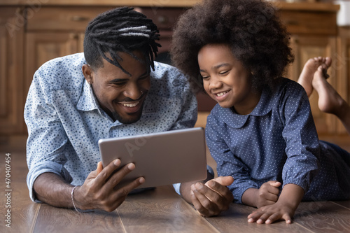 Happy young laughing African American father using digital computer tablet with little cute multiracial kid daughter, playing games online, lying together on heated floor, tech addiction concept.