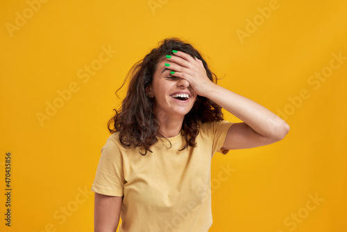 Portrait of woman with curly dark hair forgetting something, slapping forehead with palm and closing eyes isolated over yellow background