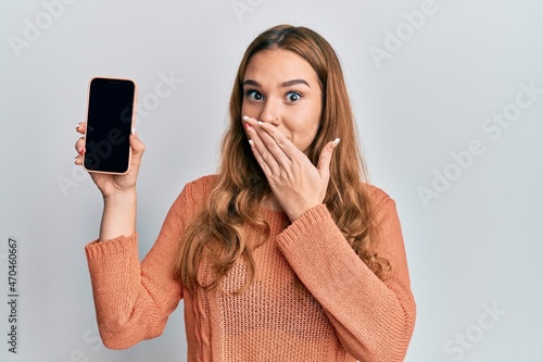 Young blonde woman holding smartphone showing screen covering mouth with hand, shocked and afraid for mistake. surprised expression
