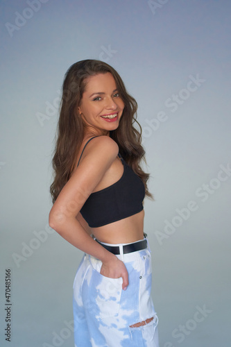 Elegant woman with natural make-up and wavy hair. Pretty girl in black top and white blue jeans with black belt posing against blue background in studio