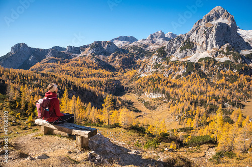 Female hiker on a bench enjoying mountain panorama view on a sunny autumn day with golden larch trees.