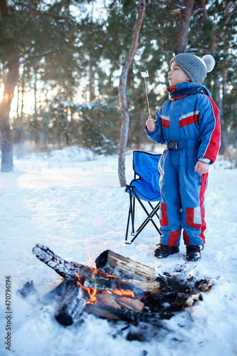 Happy boy standing by the fire in snow-covered forest and fries marshmallows, winter trip at weekend. Family winter picnic, active lifestyle