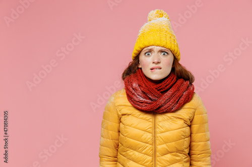 Impressed amazed panicked pop-eyed frightened scared young woman 20s years old wear ellow jacket hat mittens looking camera biting lips isolated on plain pastel light pink background studio portrait