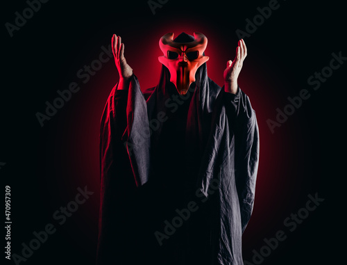 Scary horror occult sectarian priest in black hood and metal mask on black background with red glow.