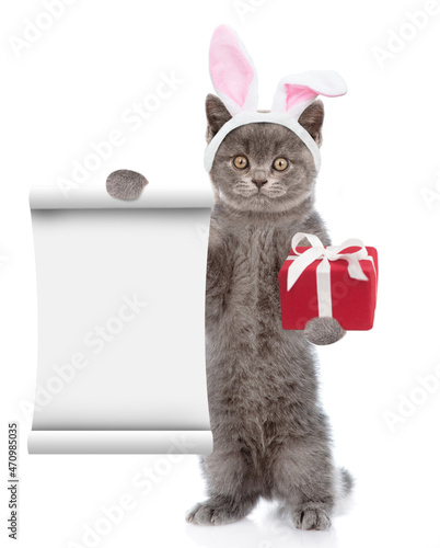 Funny kitten wearing Easter rabbits ears holds gift box end empty list. Isolated on white background