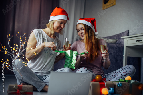 Two cute girlfriends open Christmas gift boxes during online video conference with family. Sisters exchange gifts in cozy home interior with festive decorations.