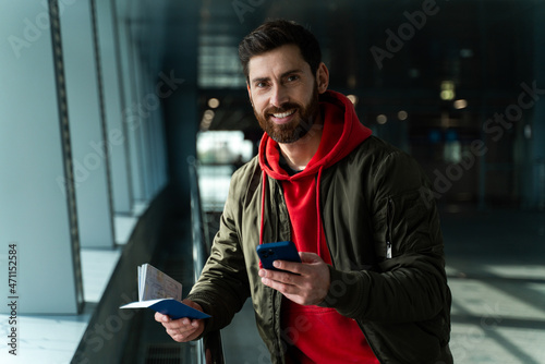 Waist up portrait of concentrated young man standing at the airport and holding his smartphone while waiting for departure. Technologies and people concept