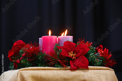 A beautiful red and green imitation Christmas wreath decoration with tinsel and poinsettia flowers encircling large lit candles and flame. Xmas concept on a simple hessian table cloth.
