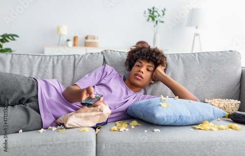 Lazy black teen guy lying on sofa with scattered food, holding TV controller, watching dull movie on television at home
