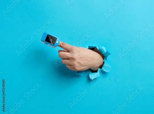 an oximeter for measuring oxygen in the blood is worn on the index finger, part of the hand is sticking out of a torn hole in the blue paper